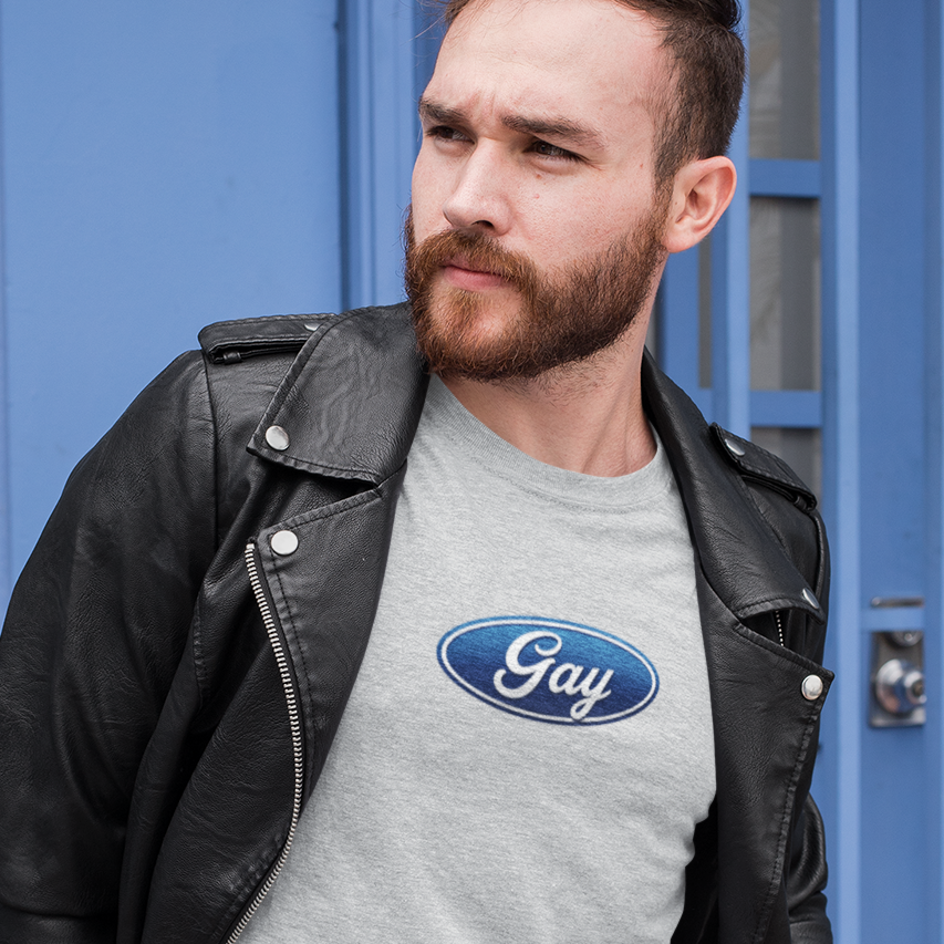A man wearing a light gray t-shirt with a blue oval 'Gay' logo, styled with a black leather jacket, standing in front of a blue door and a vintage green scooter.