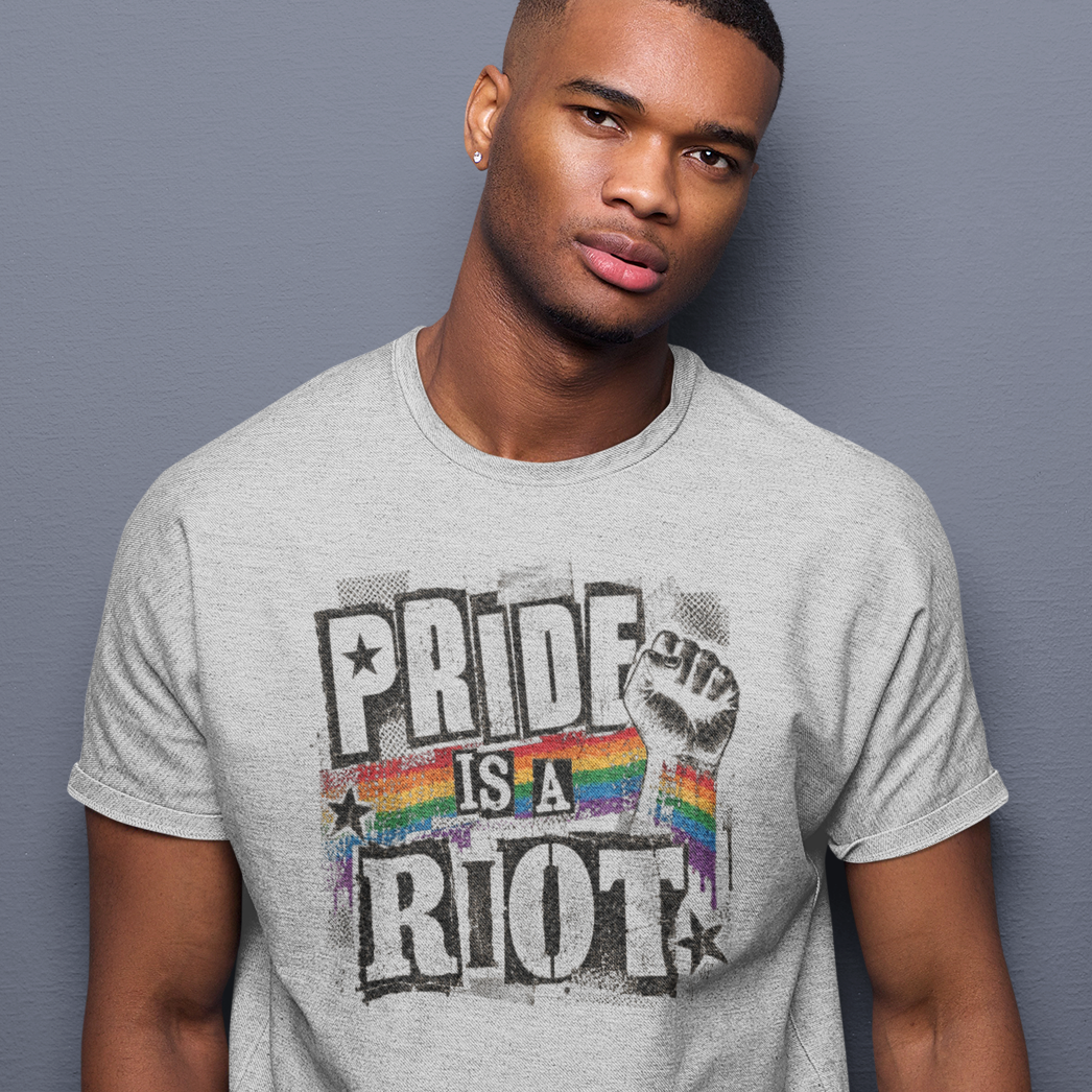 A man wearing a light gray crewneck t-shirt with a distressed graphic that reads 'PRIDE IS A RIOT' over a rainbow pride flag, posed against a solid gray background.