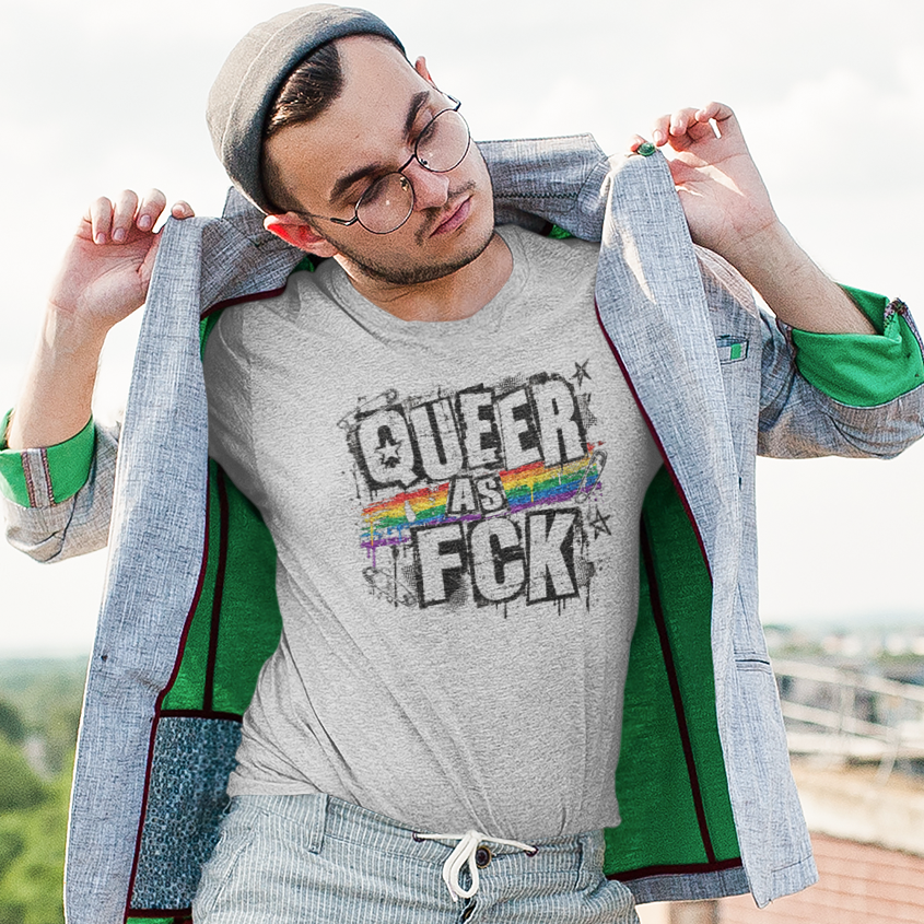 A man wearing a gray graphic t-shirt with 'QUEER AS F*CK' printed on the front, styled with a gray blazer, light gray jeans, green socks, and glasses, posing outdoors on a rooftop with a city background.