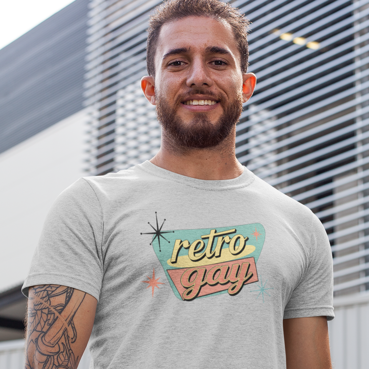 A man wearing a light gray heather t-shirt with a retro 'retro gay' graphic, standing in front of a modern building with horizontal slats.