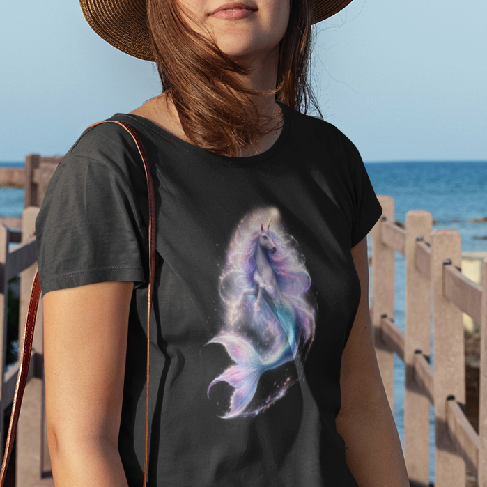 A woman stands on a wooden boardwalk by the ocean wearing a black graphic t-shirt with a purple and blue Unicorn mermaid print, denim shorts, a straw hat, and carrying a round straw bag.
