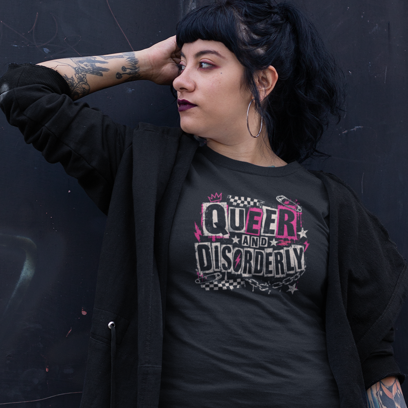 A person posing in front of a dark metal door, wearing a black graphic t-shirt that reads 'QUEER DISCOVERED', a black open-front cardigan, and a red and navy plaid mini skirt.