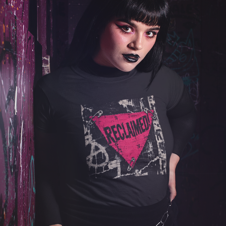 A person with black hair and black makeup stands against a graffiti-covered wall wearing a black long-sleeve t-shirt with a pink and white graphic that reads 'RECLAIMED'.
