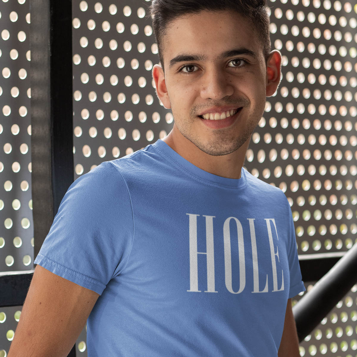 A smiling man wearing a light blue crewneck t-shirt with the word 'HOLE' printed in white across the chest, standing in front of a perforated metal wall.