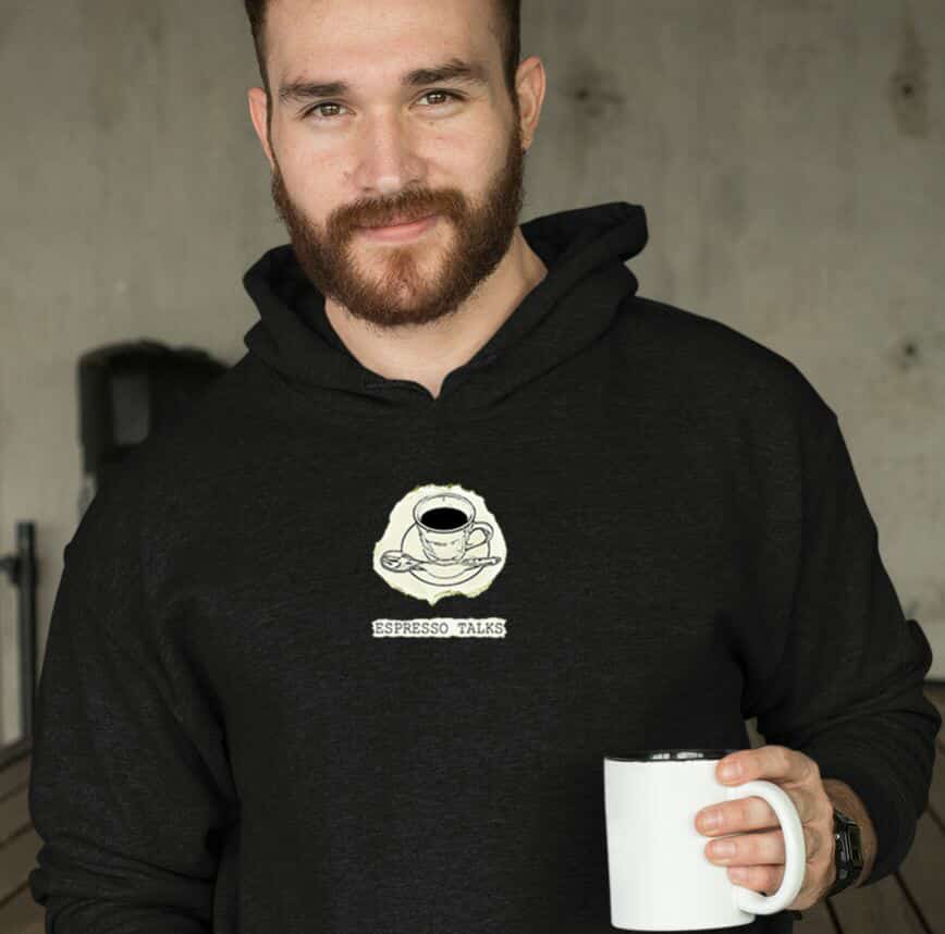 Man wearing a black hoodie with a coffee-themed design and espresso talks logo from Anthony Eaton, holding a white mug.