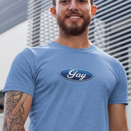 A man wearing a heathered blue t-shirt with a blue oval 'Gay' logo on the chest, standing outdoors in front of a modern slatted building wall.