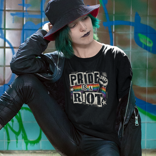 A person with green hair and gothic makeup sits on a tiled ledge in front of a graffiti wall, wearing a black bucket hat, black leather-look pants, a black graphic T-shirt that reads 'PRIDE OR RIOT', black knee-high socks, and black boots.