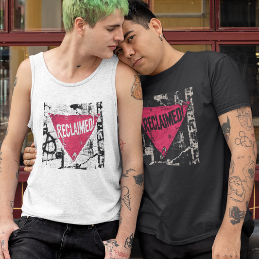 Two men standing outdoors in front of a building with red-framed windows. The man on the left has bright green hair and wears a white tank top with a pink and black graphic that reads 'RECLAIMED'.