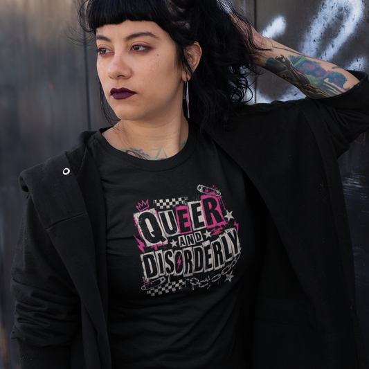A person with dark hair and tattoos stands against a dark metal wall wearing a black graphic T-shirt that reads 'QUEER AND DISORDERLY' in pink, white, and black, layered under an open black coat, with a plaid mini skirt and fishnet stockings.